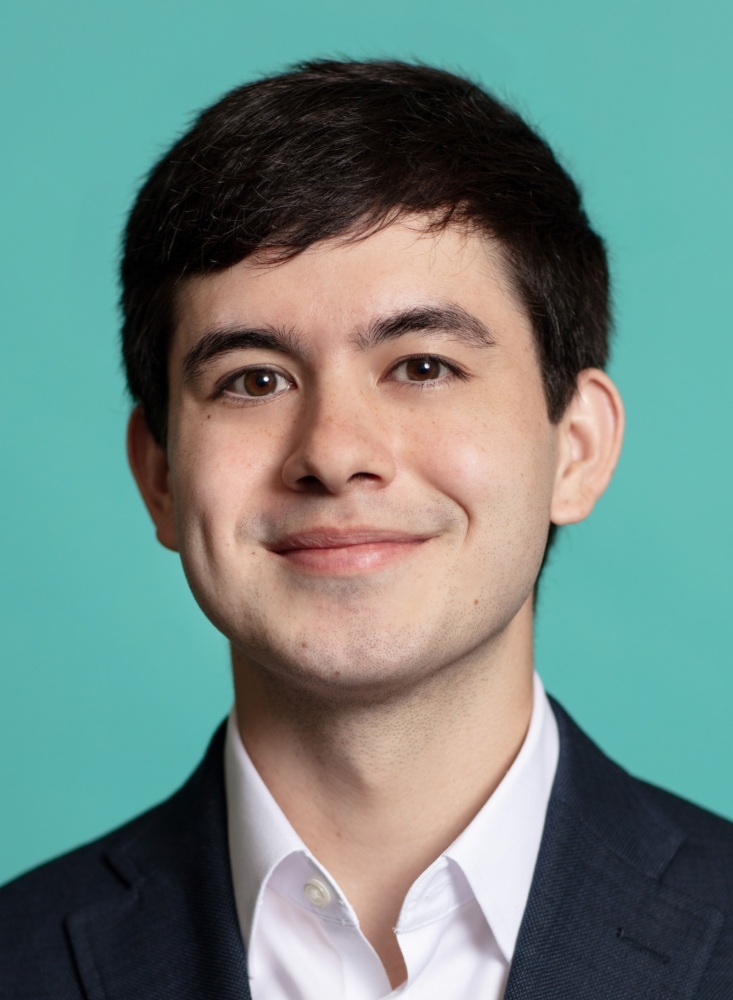 Portrait of a young man with short dark hair wearing a dark suit jacket and white shirt, standing against a solid teal background. He is looking at the camera with a slight smile.